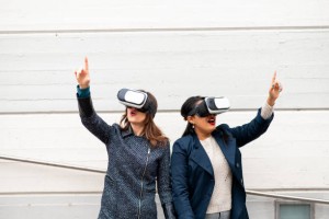 Two attractive young women leaning shoulder to shoulder wearing virtual reality headsets. Females with surprised faces pointing something.