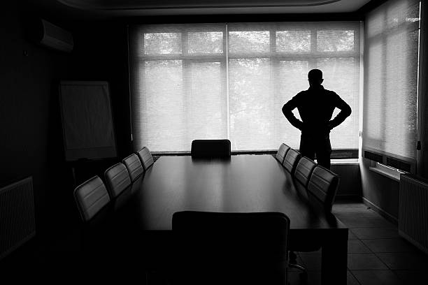 Silhouette Of Lonely Businessman Standing By Boardroom Table In Office.The chairs around table are empty.He looks desperate.The black and white color and dark atmosphere is used for economic depression feel.Full frame DSLR camera was used in real office.