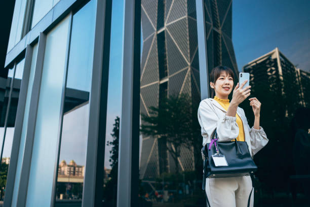 This is a photo of a modern and successful young Asian businesswoman carrying a smartphone and a business bag, commuting to work in the central business district of the city. The background consists of contemporary corporate buildings, and she is seen using a mobile device for communication, showcasing her profession. Her expression exudes passion and focus towards her work.
